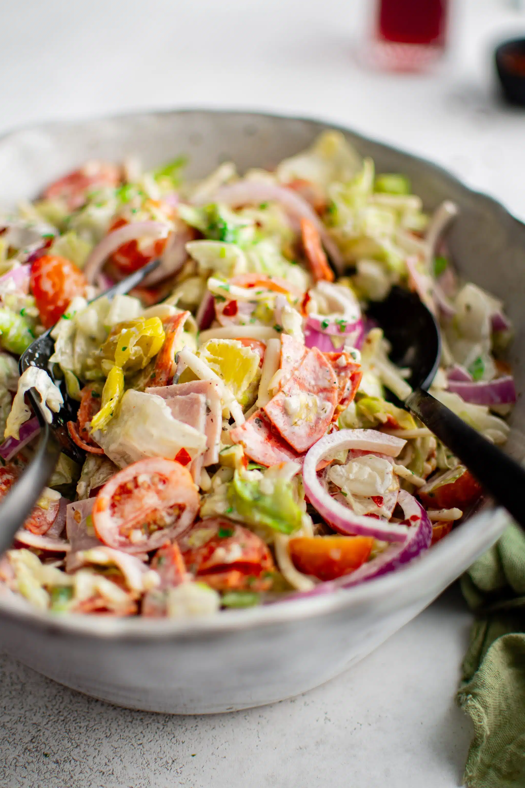 Grinder salad presented in a large white ceramic salad bowl filled with chopped iceberg lettuce, halved cherry tomatoes, sliced red onion, sliced pepperoncini peppers, and sliced ham, pepperoni, and salami.