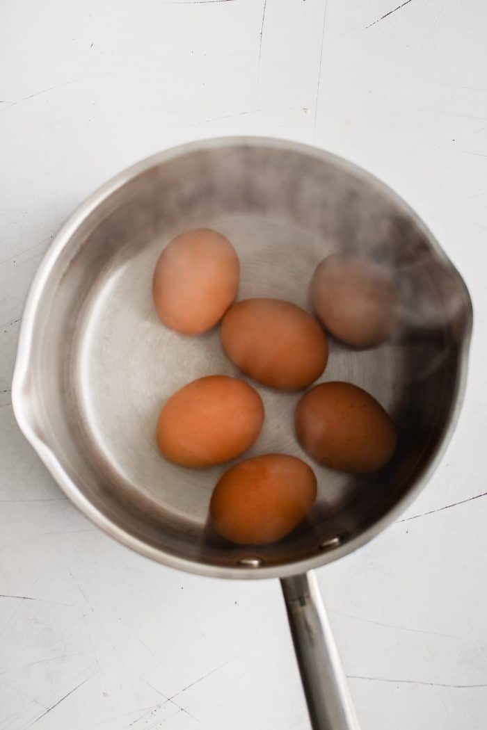 An overhead view of six brown eggs gently boiling in a stainless steel saucepan filled with water. 