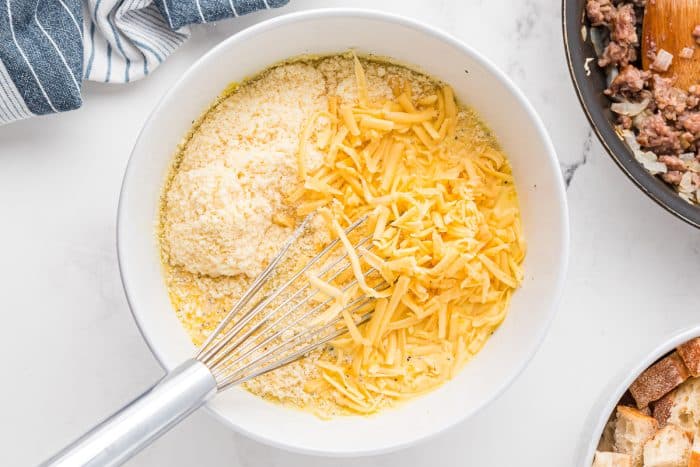 Shredded gouda and grated parmesan added to a bowl of whisked eggs and milk, showing the next step in making a bread-based breakfast casserole.