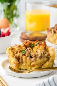 A golden slice of breakfast casserole served on a white plate with a fork, garnished with fresh parsley and paired with orange juice.