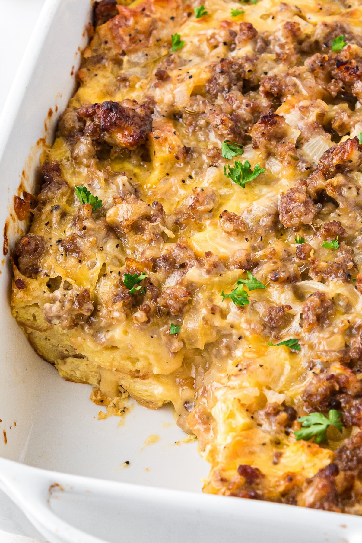 Close-up of baked breakfast casserole with golden bread cubes, melted cheese, browned sausage, and parsley garnish.