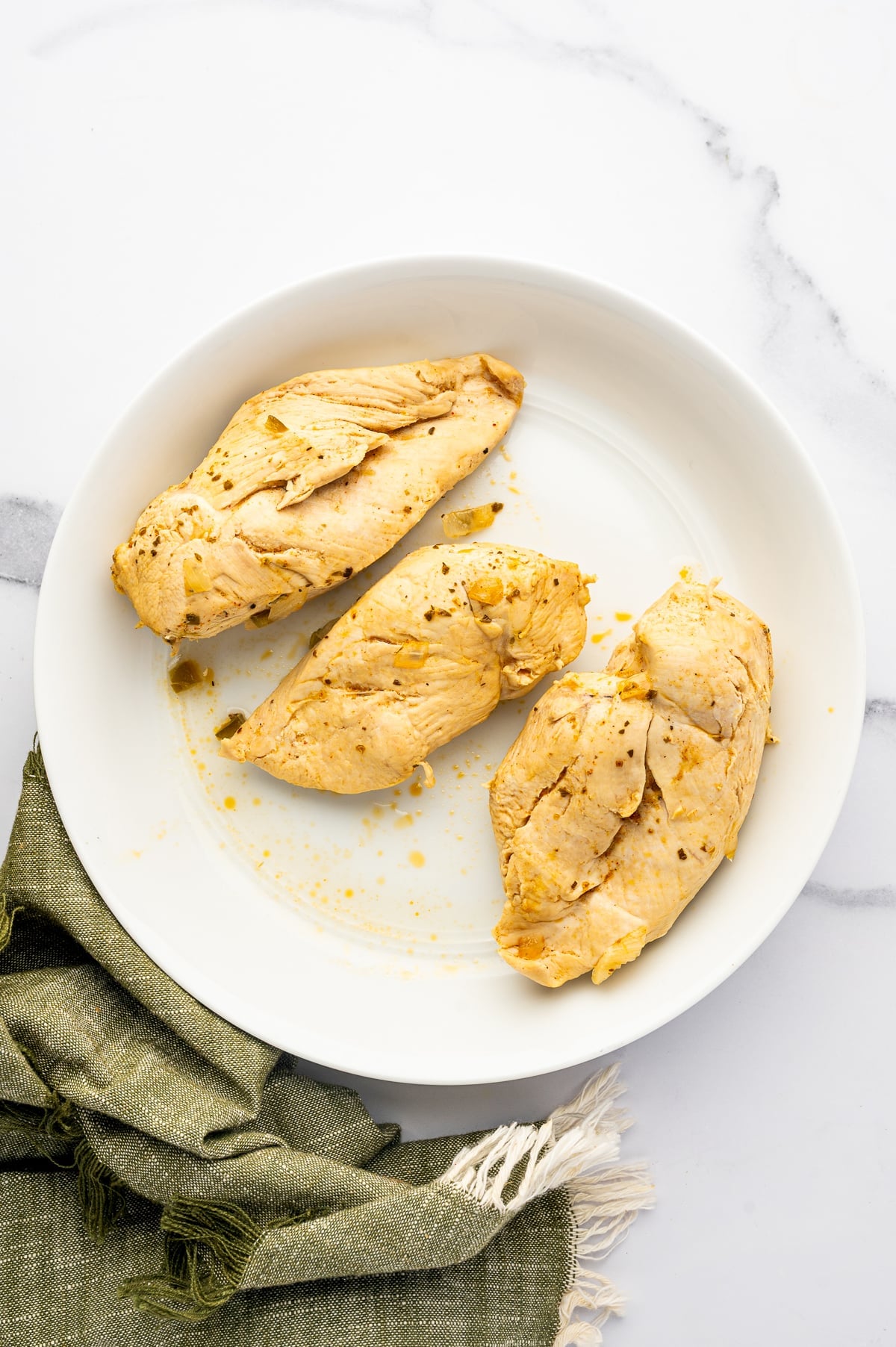 Three cooked, lightly seasoned chicken breasts resting on a large white plate beside an olive-green kitchen towel after being removed from the chili broth.