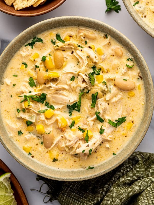 Close overhead view of creamy white bean chicken chili in a speckled sage-green bowl filled with shredded chicken, whole cannellini beans, yellow corn, green chiles, and chopped fresh cilantro.