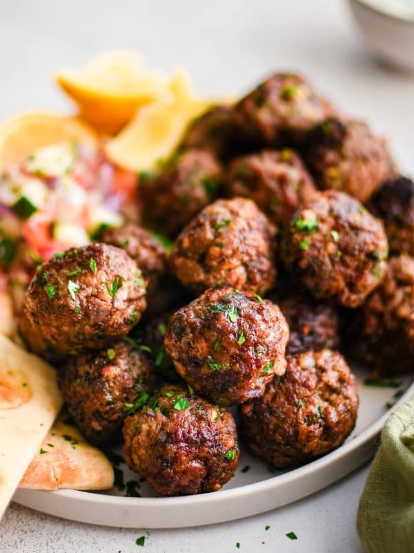 Close-up side view of browned Greek meatballs garnished with chopped parsley on a light beige plate with pita bread, lemon wedges, a blurred bowl of tzatziki, and a green cloth napkin on a white textured surface.