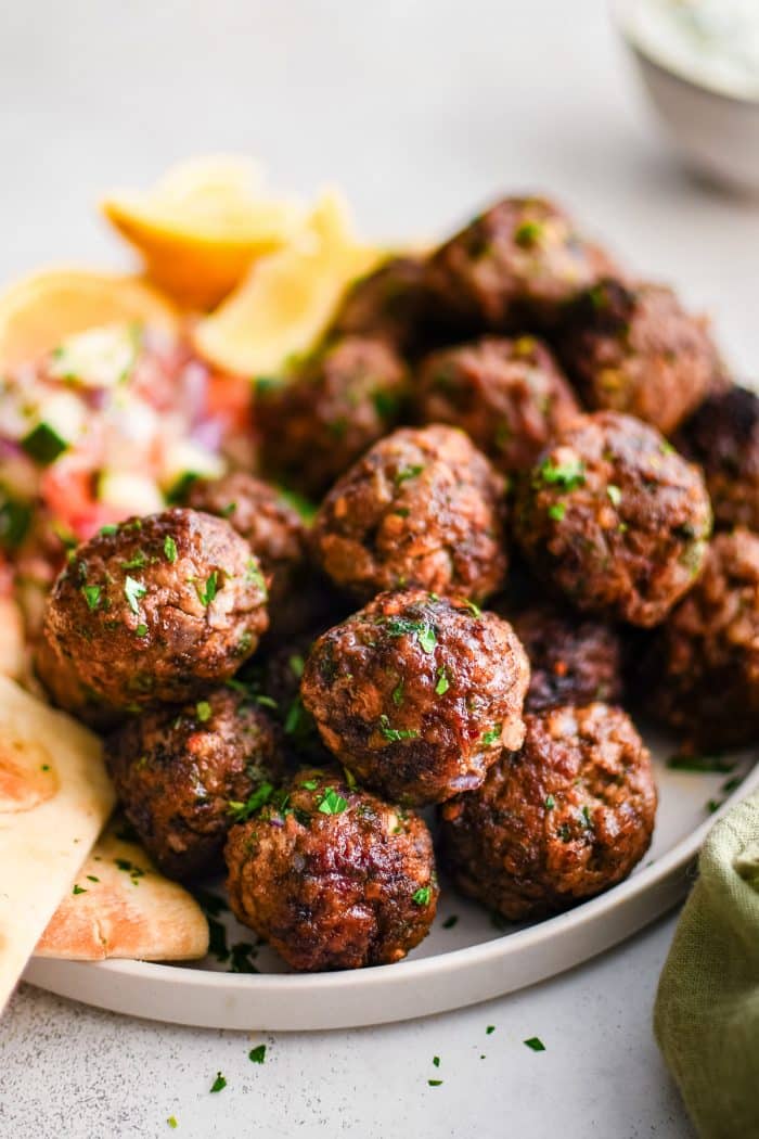 Close-up side view of browned Greek meatballs garnished with chopped parsley on a light beige plate with pita bread, lemon wedges, a blurred bowl of tzatziki, and a green cloth napkin on a white textured surface.