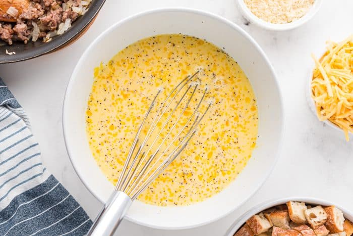 Large white mixing bowl filled with whisked eggs, milk, and seasonings with a metal whisk, ready to be combined for an easy breakfast casserole.