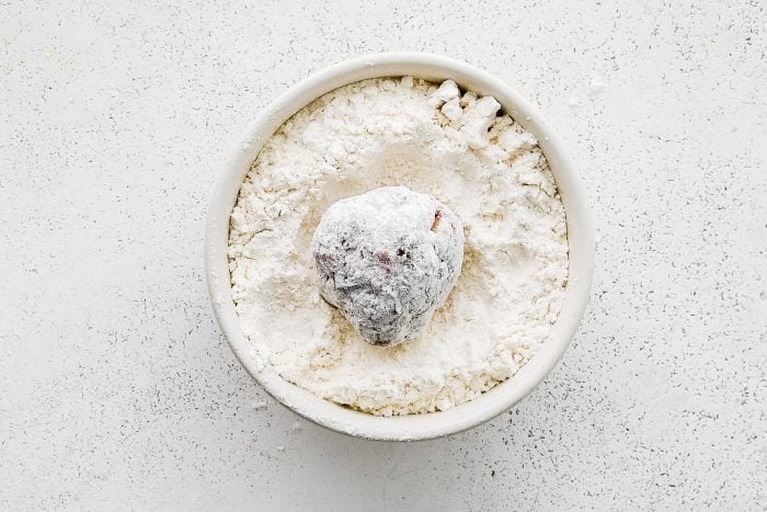 Close-up overhead view of one raw Greek meatball coated in white flour inside a small white bowl filled with flour on a white textured surface.