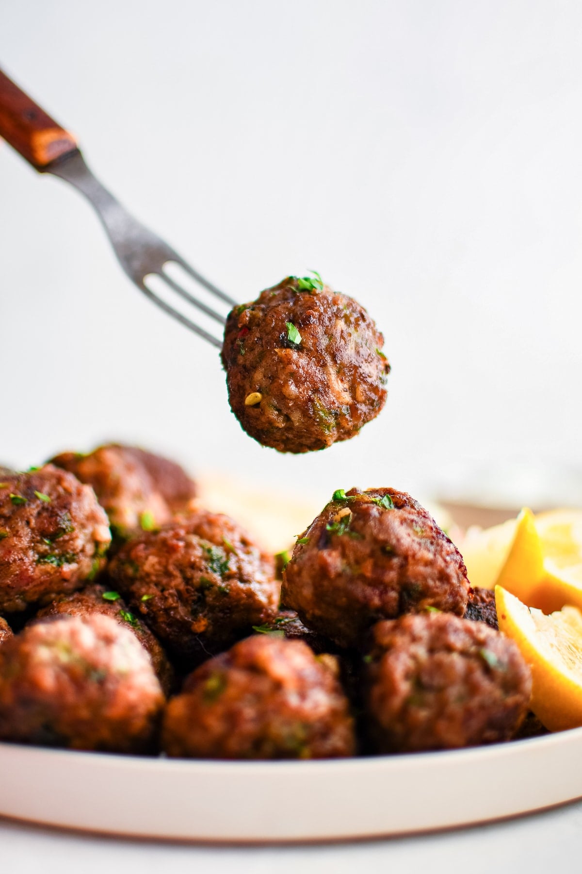 Close-up of one golden brown Greek meatball lifted on a fork over a plate of meatballs with chopped parsley, lemon wedges, and a softly blurred white background.