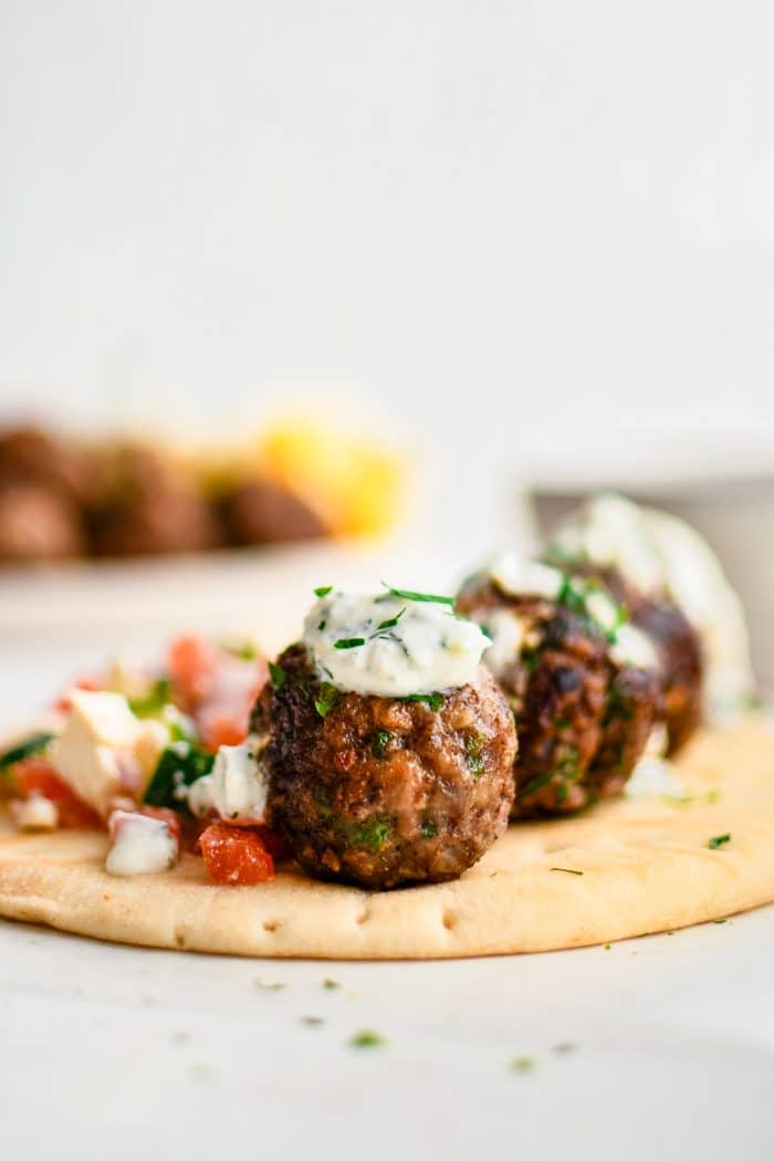 Close-up of a browned Greek meatball topped with creamy tzatziki sauce and chopped parsley on soft pita bread, with chopped tomato cucumber salad and another meatball in the background.