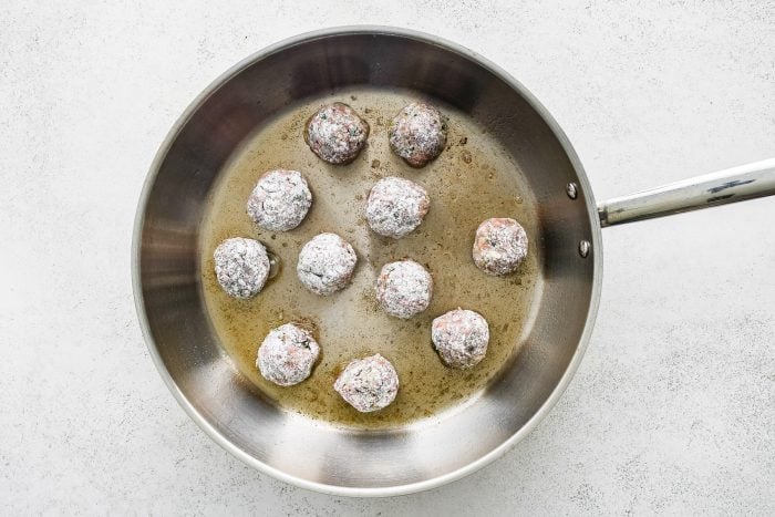 Overhead view of flour-coated raw Greek meatballs cooking in a large stainless steel skillet with oil on a white textured surface.