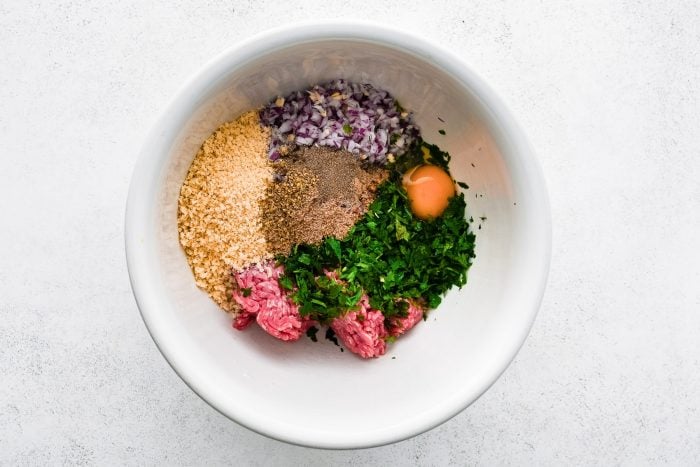 Overhead view of Greek meatball ingredients in a large white mixing bowl, including ground beef, chopped fresh herbs, minced red onion and garlic, panko breadcrumbs, an egg, and seasonings on a white textured surface.