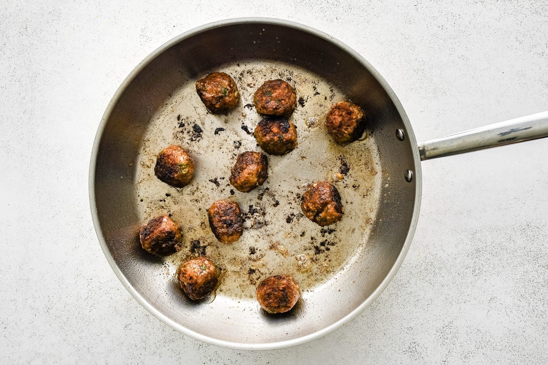 Overhead view of browned Greek meatballs cooking in a large stainless steel skillet with oil and dark browned bits on the bottom of the pan on a white textured surface.