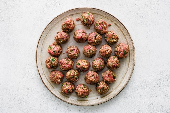 Overhead view of raw Greek meatballs rolled into small balls and arranged on a beige oval plate with a brown rim on a white textured surface.
