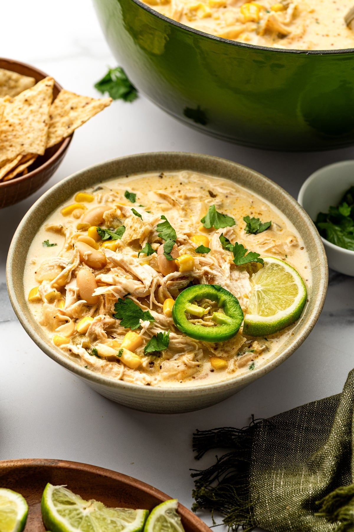 Creamy white chicken chili served in a speckled sage-green ceramic bowl topped with cilantro, a jalapeno slice, and lime, with a dark green enameled Dutch oven and tortilla chips in the background.