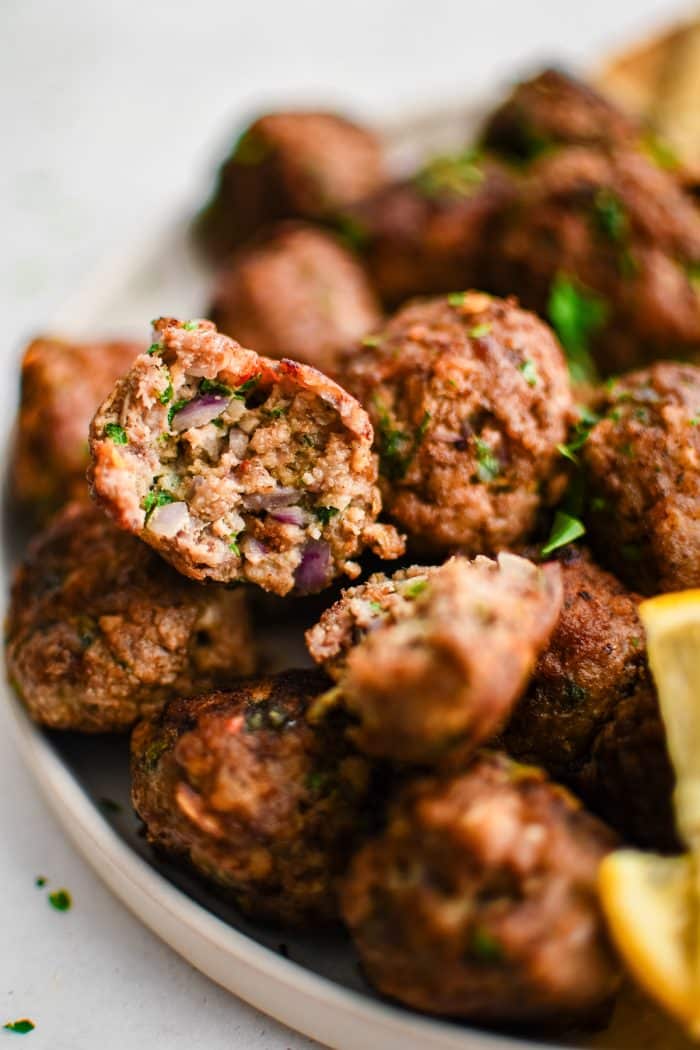 Close-up of a sliced Greek meatball showing the juicy interior with bits of red onion, breadcrumbs, and fresh herbs, served over a plate of browned meatballs with chopped parsley and lemon wedges.