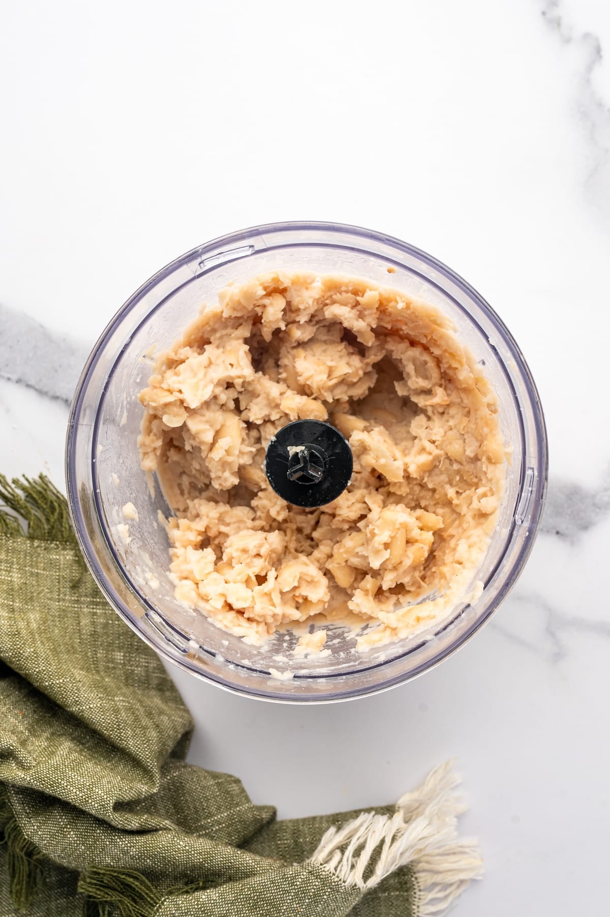 Overhead view of drained cannellini beans processed into a thick, pale beige puree inside a small clear food processor on a white marble surface.