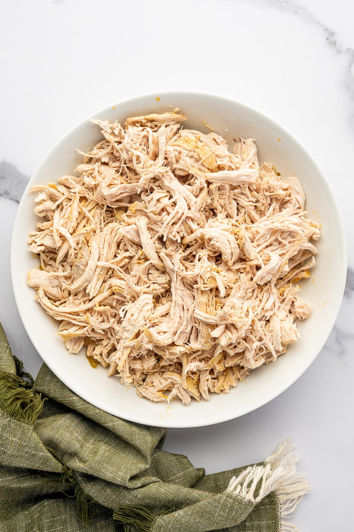 Overhead view of a large pile of finely shredded cooked chicken on a round white plate with an olive-green cloth partially visible along the lower edge.