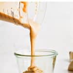 Creamy homemade tahini paste being poured from a measuring cup into a clear glass jar, with lemon wedges in the background and the text "how to make tahini paste" at the top.