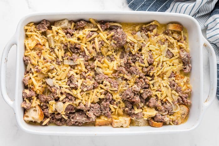 White casserole dish filled with cubed bread, cooked sausage, onions, and cheese layered together before baking an easy breakfast casserole.