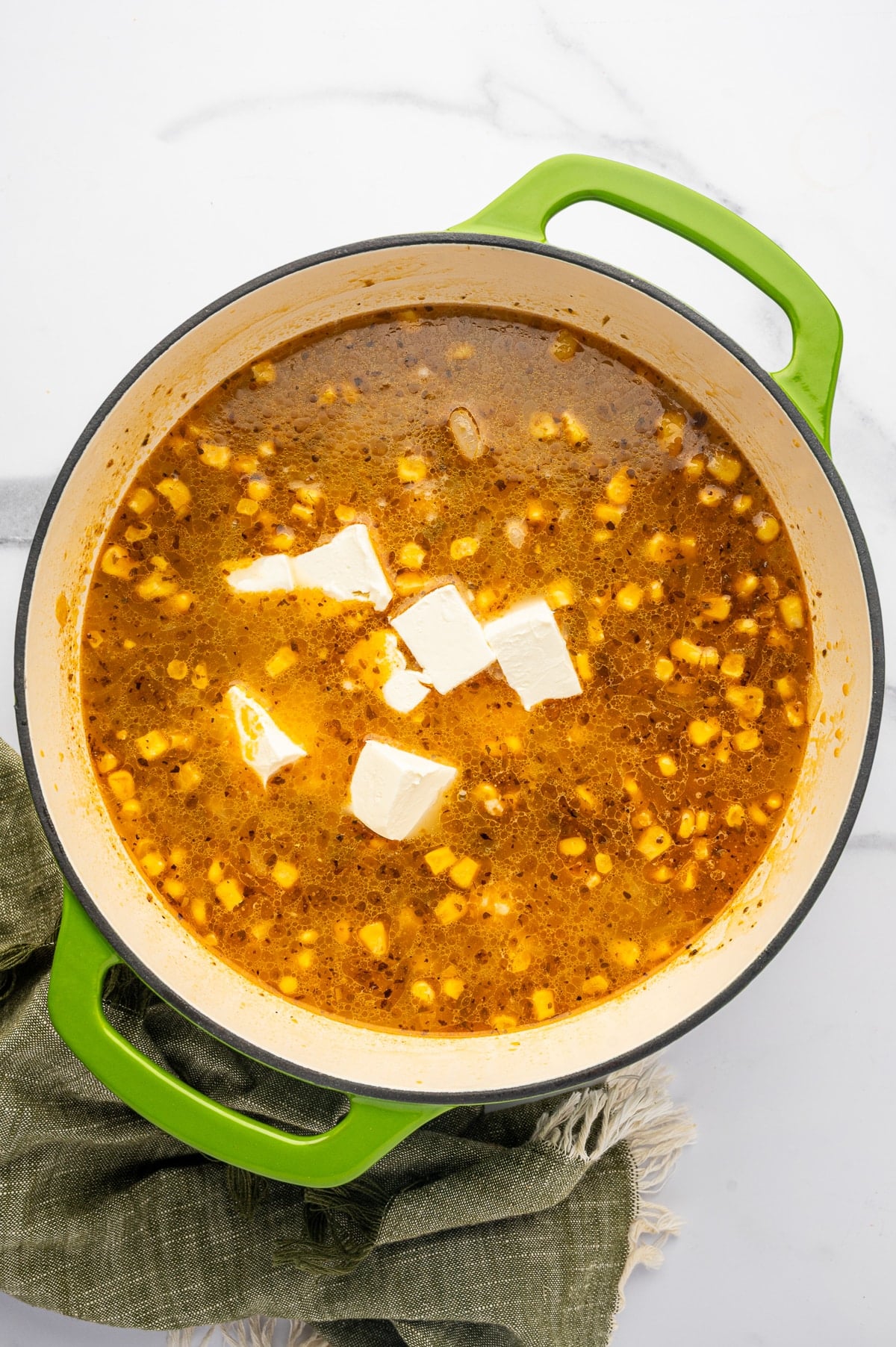 Overhead view into a lime-green Dutch oven filled with seasoned chili broth and yellow corn, with several white cubes of cream cheese floating on top before melting.
