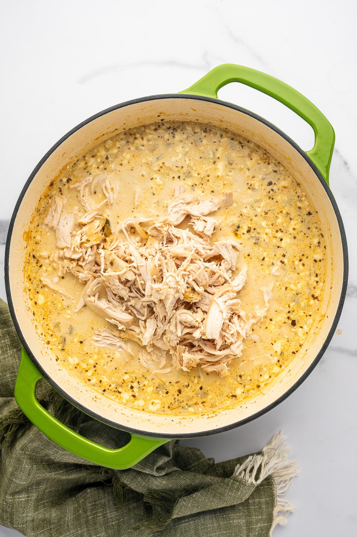 Overhead view of shredded chicken piled in the center of thick, creamy white chicken chili inside a lime-green enameled Dutch oven with visible herbs and corn.