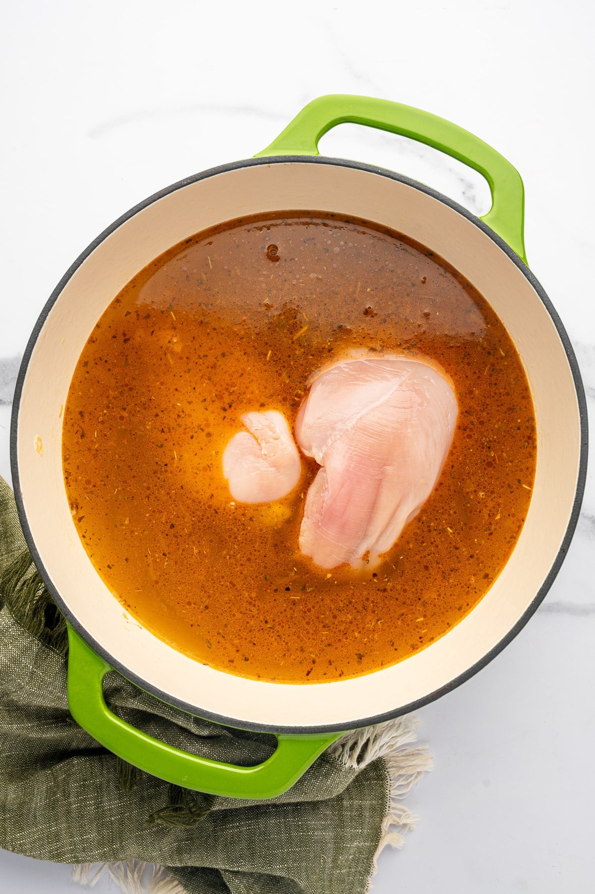 Raw boneless skinless chicken breasts submerged in reddish seasoned chicken broth inside a bright lime-green enameled Dutch oven with a cream interior.