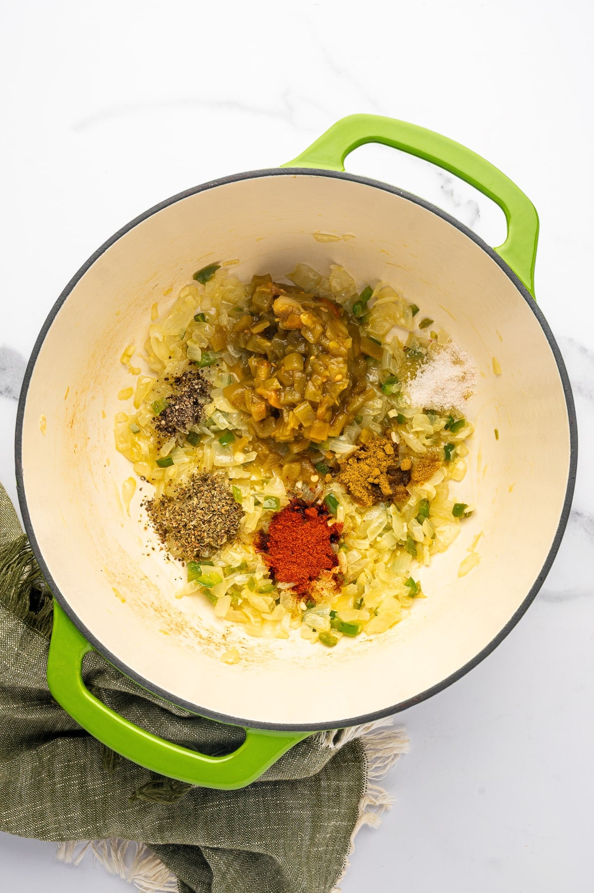 Overhead view of softened onion and jalapeno in a lime-green Dutch oven topped with diced green chiles, chili powder, cumin, dried oregano, salt, and black pepper before stirring.