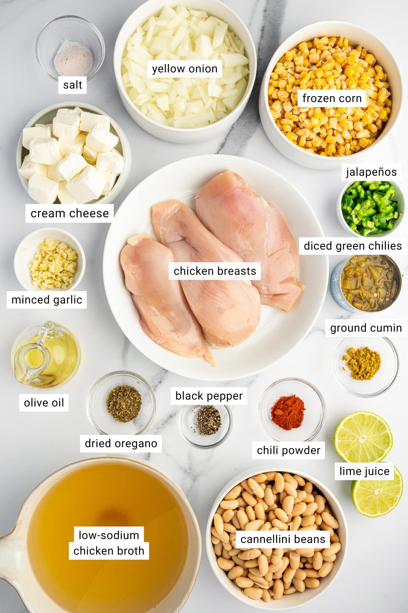 Overhead ingredient layout for white chicken chili with labeled chicken breasts, cannellini beans, frozen corn, cream cheese, yellow onion, jalapenos, diced green chiles, garlic, chicken broth, olive oil, lime juice, chili powder, cumin, oregano, salt, and black pepper.