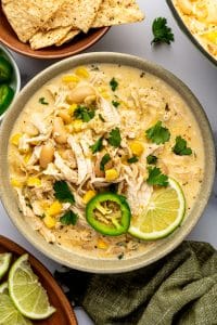 Overhead close-up of creamy white chicken chili in a speckled sage-green ceramic bowl, topped with shredded chicken, cannellini beans, corn, cilantro, a fresh jalapeno slice, and a lime wedge, with tortilla chips nearby.