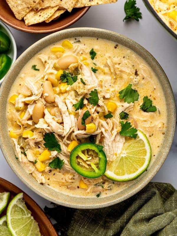 Overhead close-up of creamy white chicken chili in a speckled sage-green ceramic bowl, topped with shredded chicken, cannellini beans, corn, cilantro, a fresh jalapeno slice, and a lime wedge, with tortilla chips nearby.