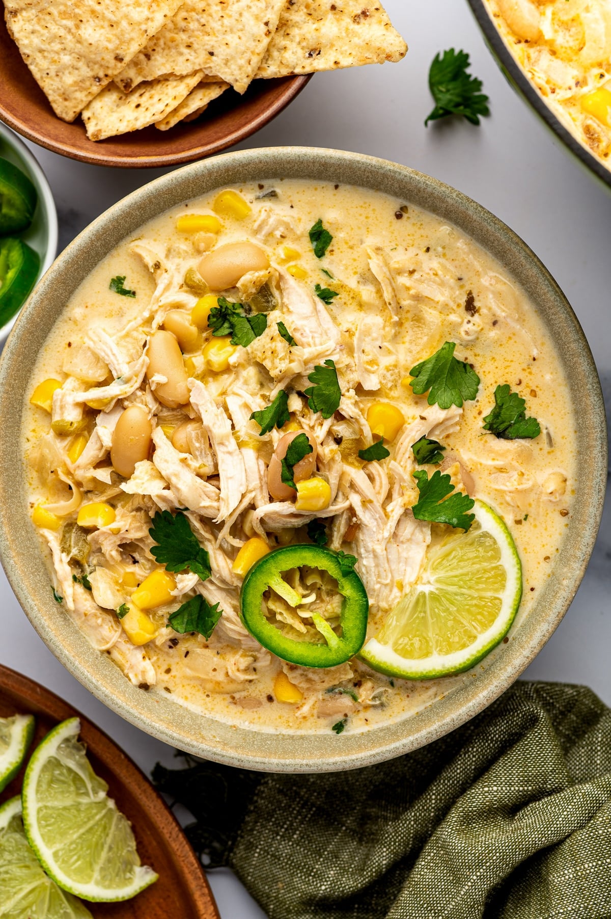 Overhead close-up of creamy white chicken chili in a speckled sage-green ceramic bowl, topped with shredded chicken, cannellini beans, corn, cilantro, a fresh jalapeno slice, and a lime wedge, with tortilla chips nearby.