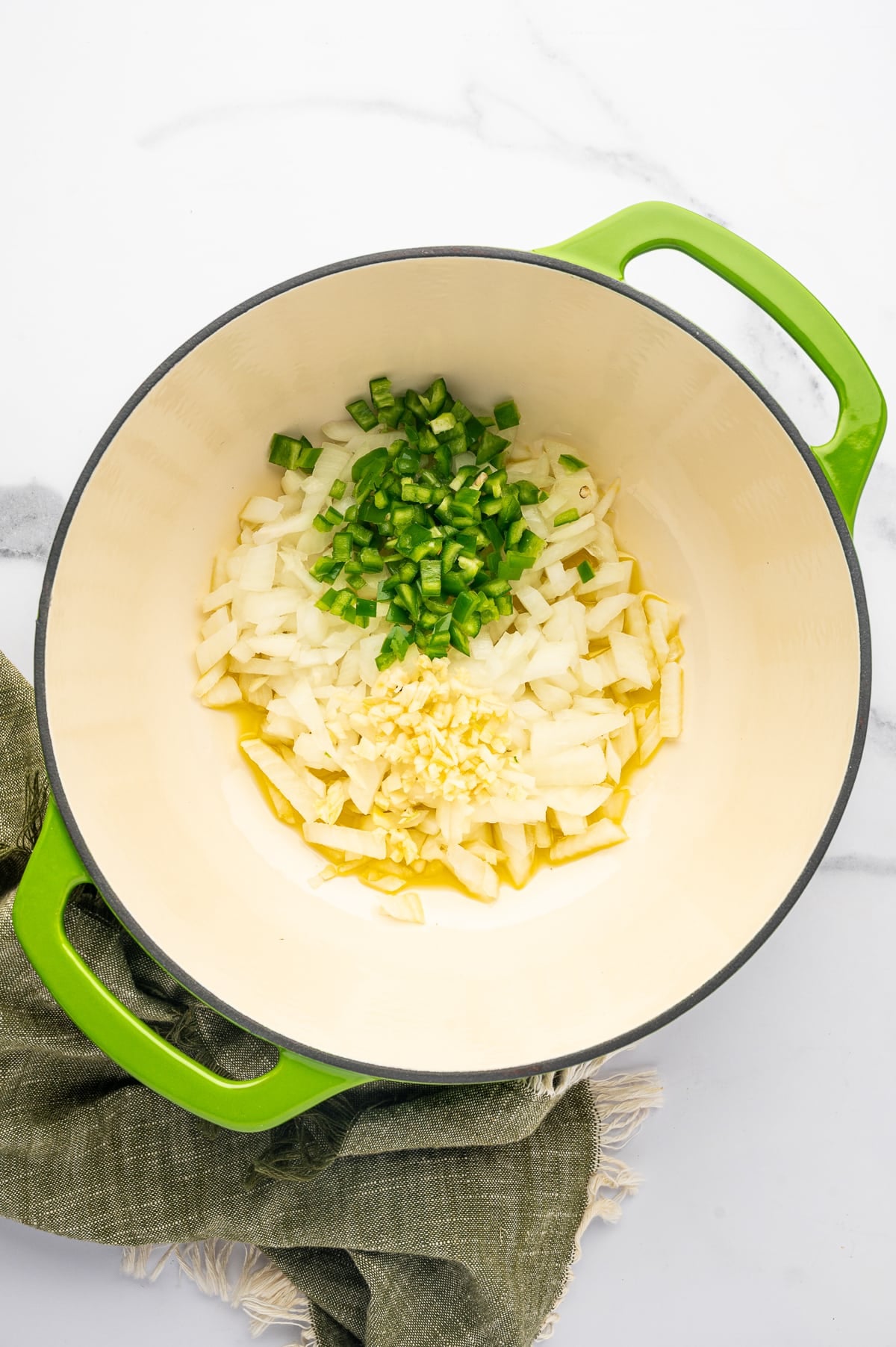 Overhead view of chopped yellow onion, diced green jalapenos, and minced garlic in olive oil inside a bright lime-green enameled Dutch oven with a cream-colored interior.