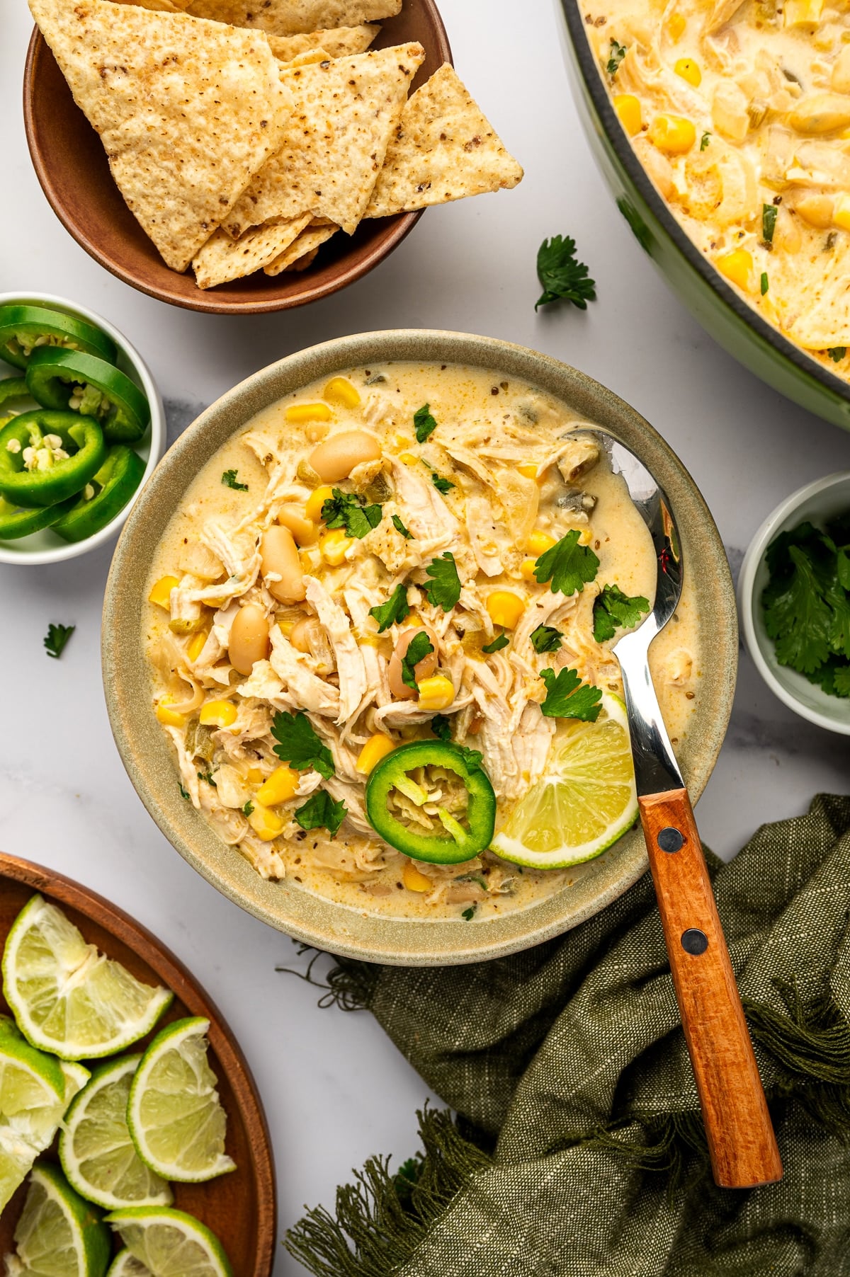 Overhead serving scene with creamy white chicken chili in a speckled sage-green bowl topped with cilantro, jalapeno, and lime, surrounded by tortilla chips, sliced jalapenos, lime wedges, and the green Dutch oven.