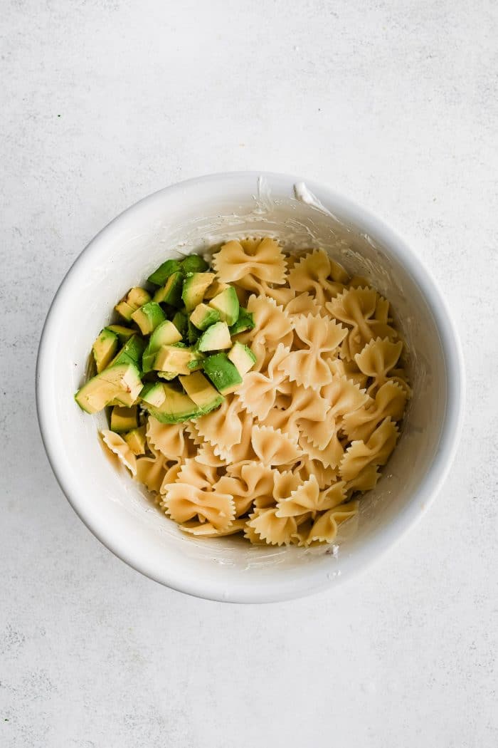 Cooked bowtie pasta and cubed avocado added to a large white bowl.