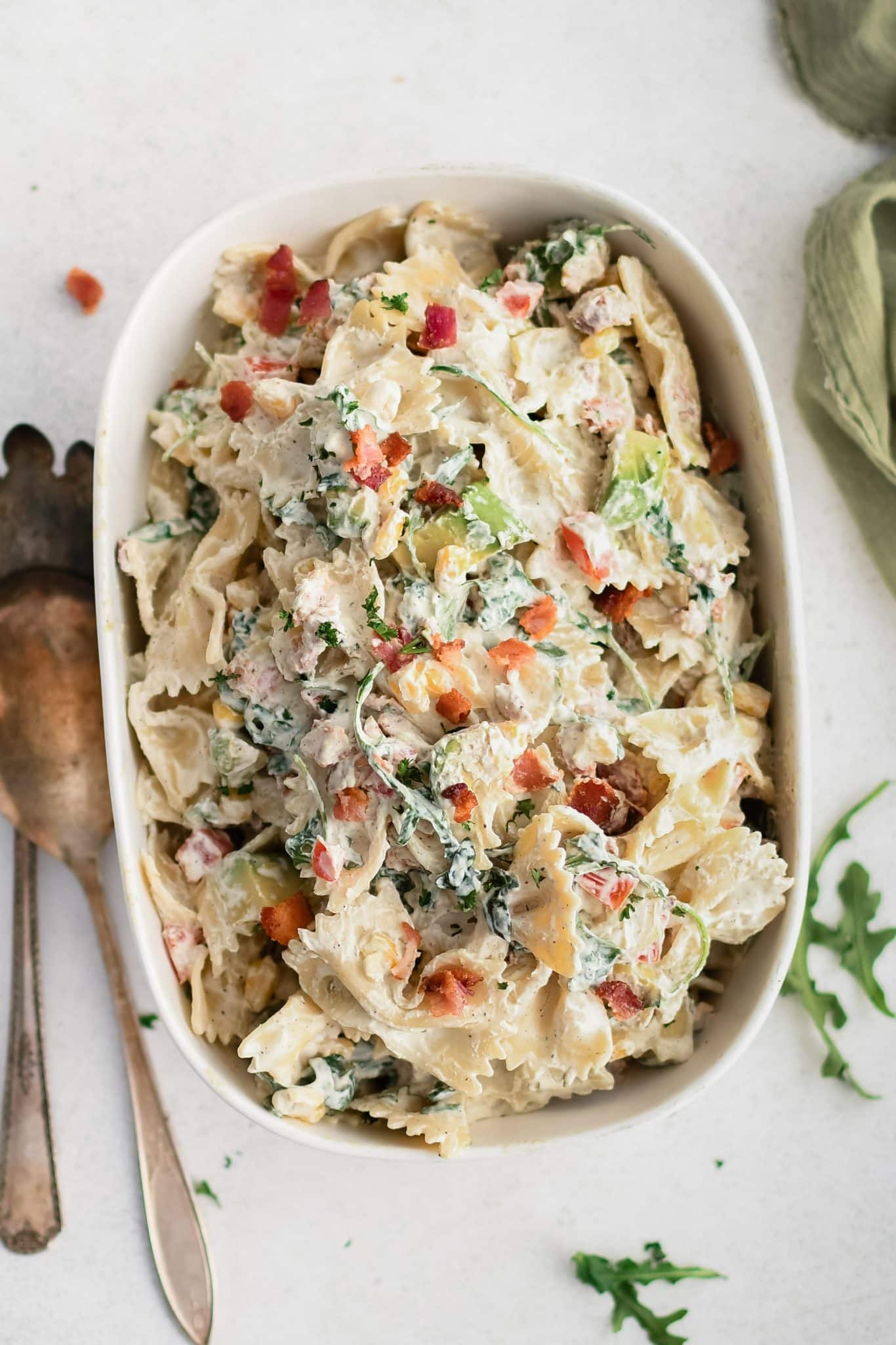 Large oval serving plate filled with creamy BLT pasta salad made with bowtie pasta, arugula, avocado, tomato, bacon, and corn in a creamy mayo and sour cream dressing.