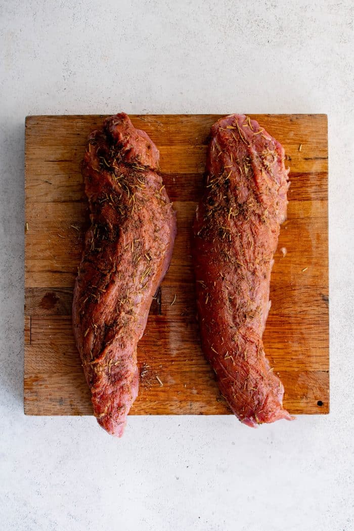 Two raw pork tenderloins on a wooden cutting board coated with a dry spice rub of paprika, rosemary, black pepper, and salt.