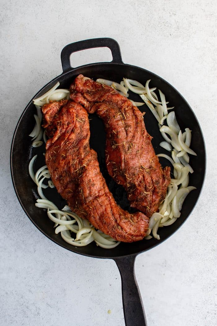 Two seasoned pork tenderloins placed on top of sautéed onions and garlic in a cast-iron skillet.