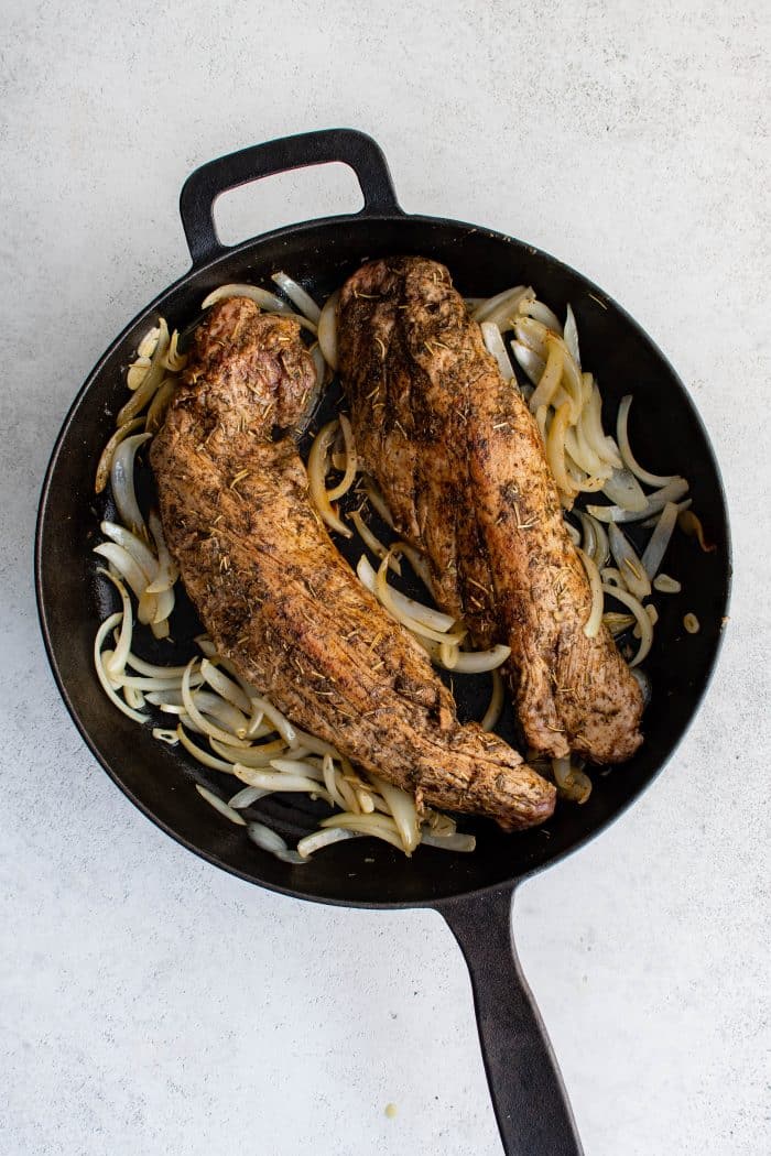 Two pork tenderloins seared until browned on the outside, resting on a bed of sautéed onions in a cast-iron skillet.