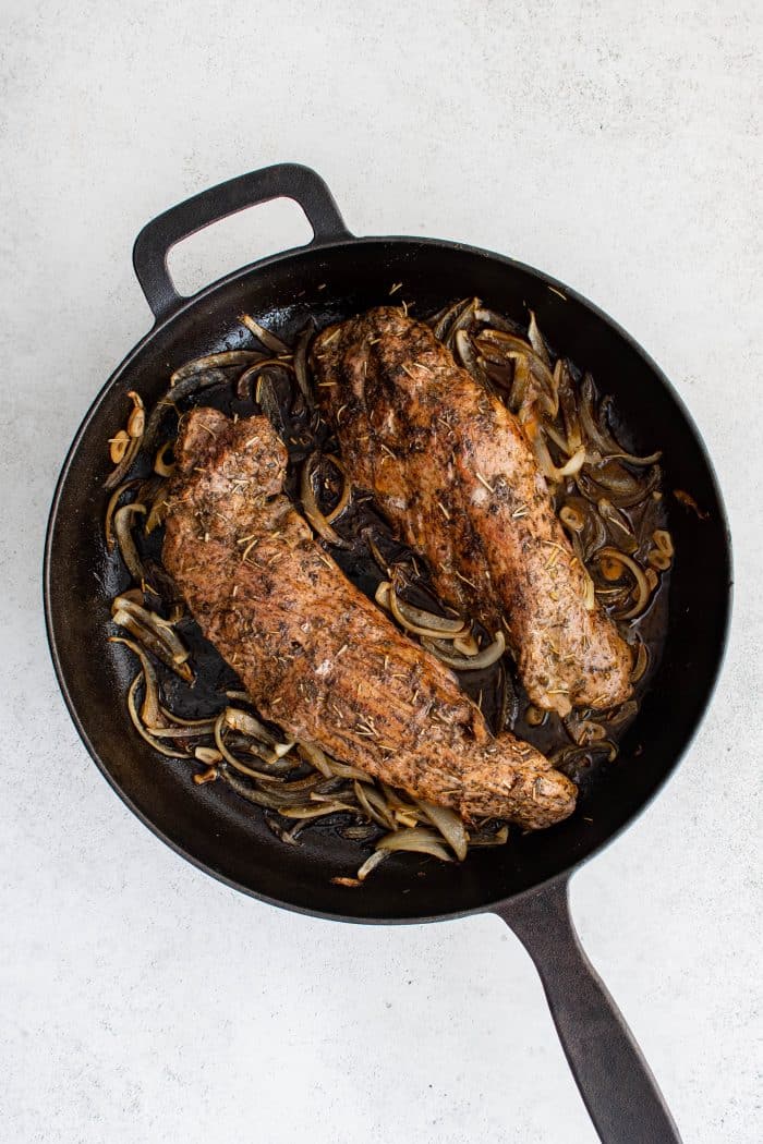 Fully cooked pork tenderloins resting in a cast-iron skillet with caramelized onions after baking in the oven.