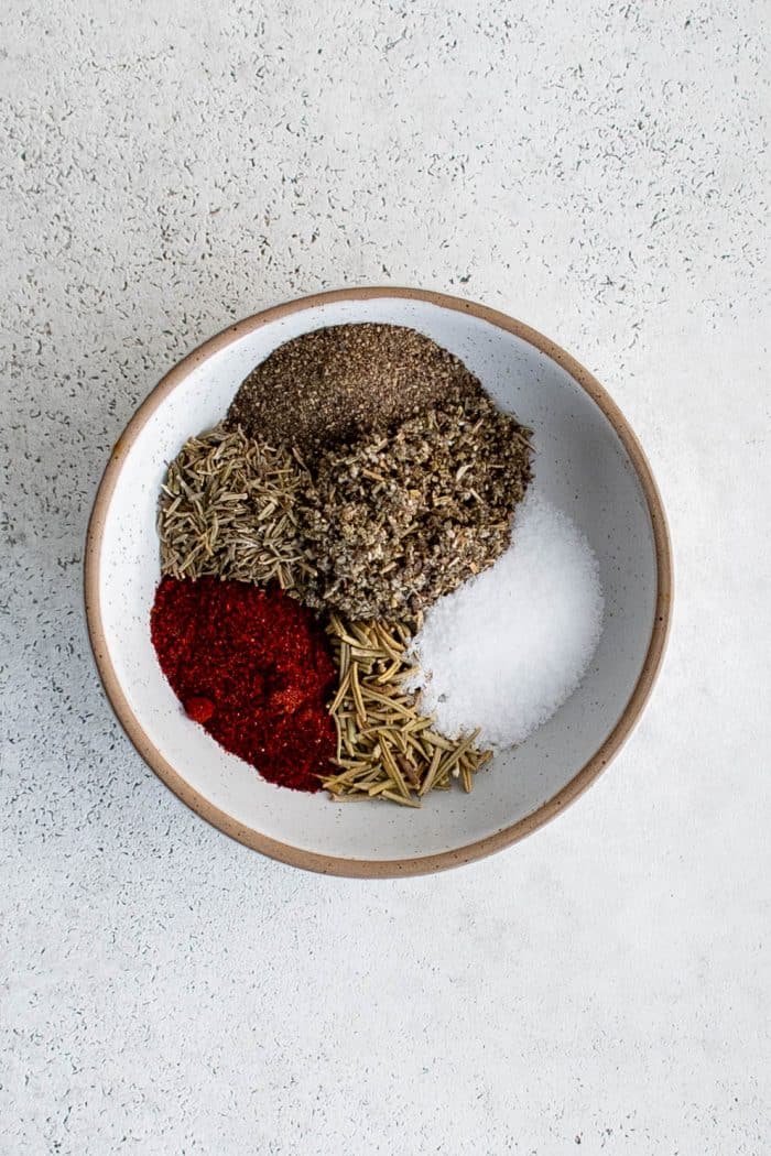 Overhead view of a white ceramic bowl filled with a homemade spice mix for pork tenderloin, including paprika, salt, black pepper, rosemary, and dried thyme.