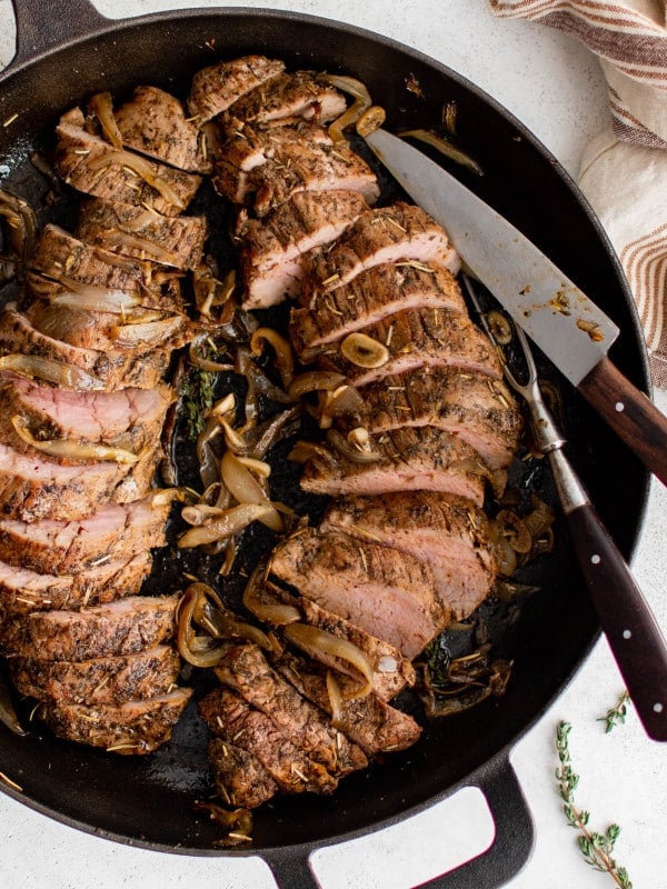 Large cast iron skillet filled with two cooked and seasoned baked pork tenderloin sliced into medallions.