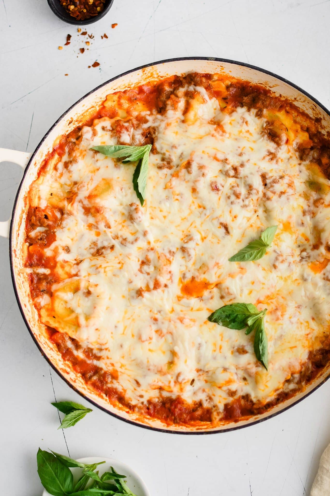 Gooey melted mozzarella cheese covering a large skillet filled with ravioli coated with meat-filled marinara sauce and garnished with fresh basil leaves.