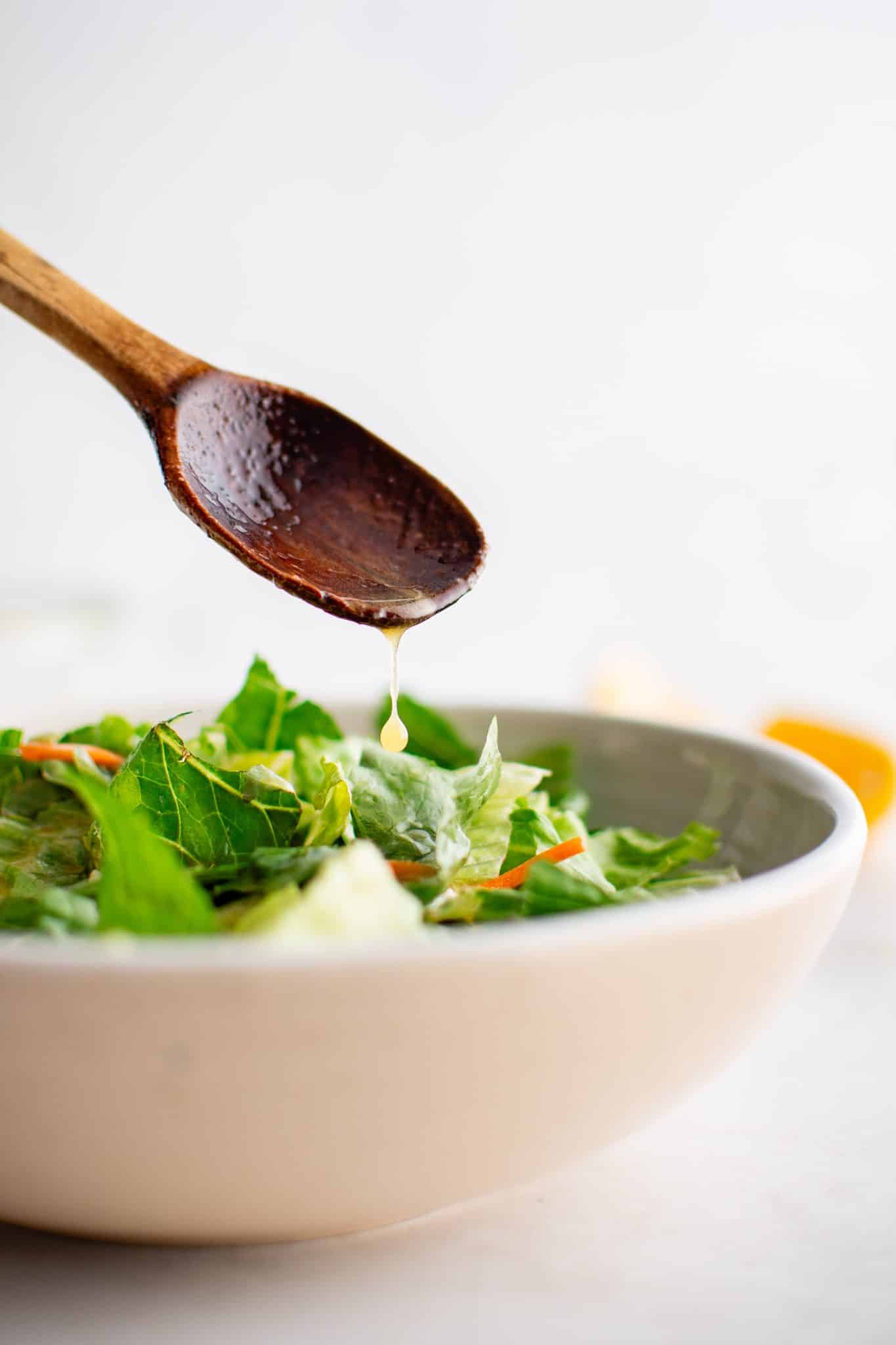 Small wood spoon drizzling homemade champagne vinaigrette over a large white bowl filled with fresh greens.