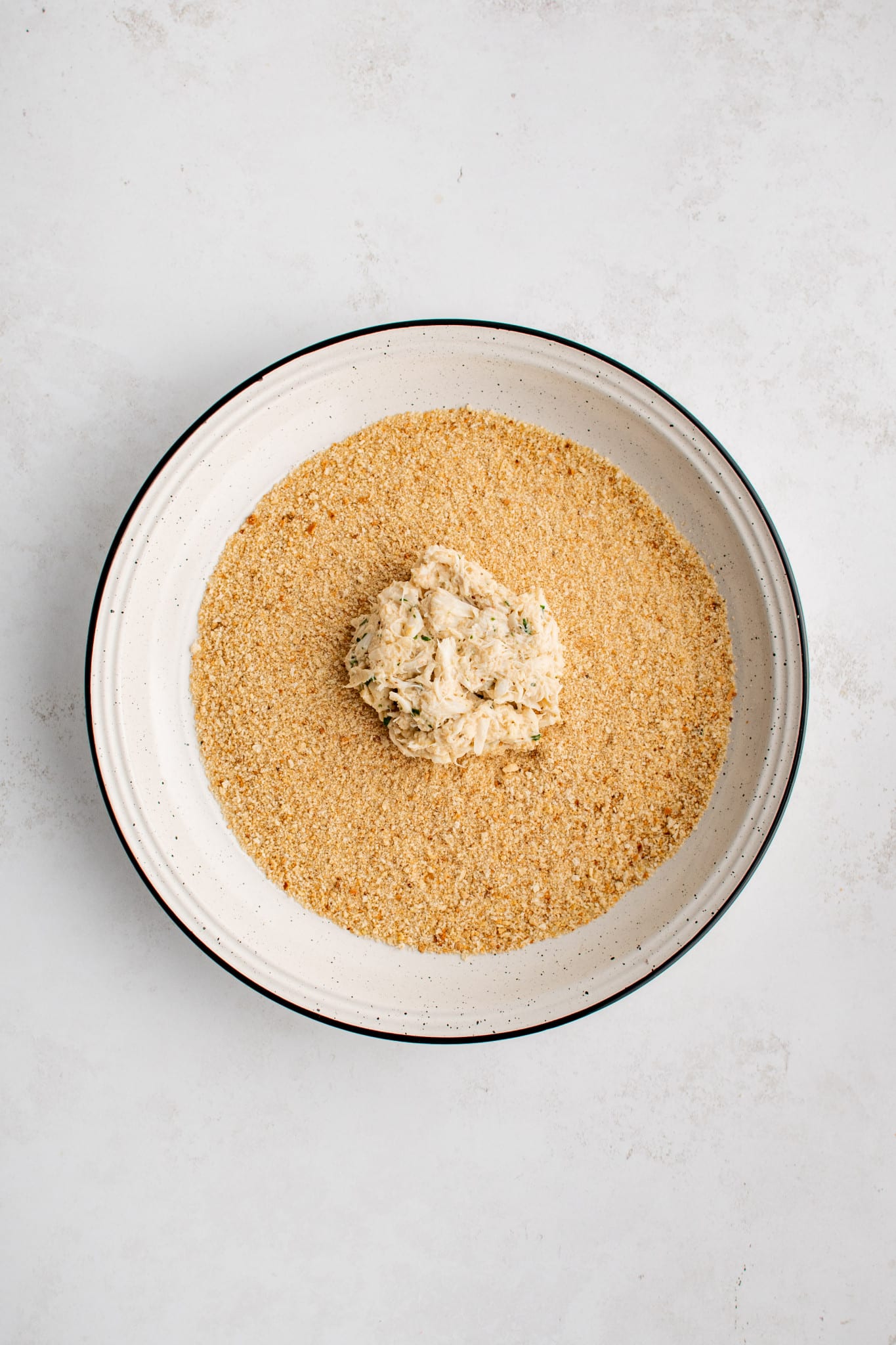 One formed crab cake in a large shallow bowl filled with plain breadcrumbs.