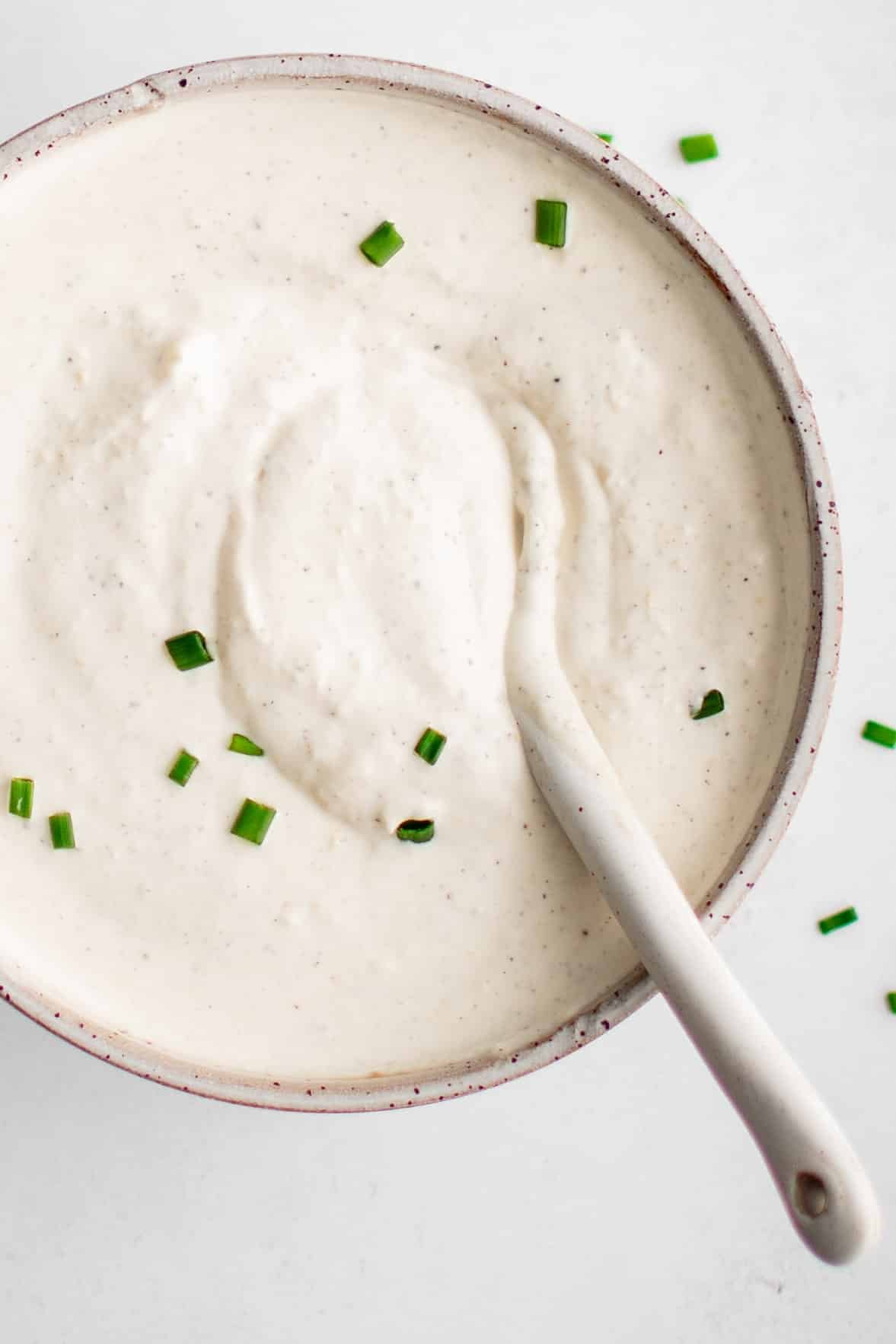 A white spoon scooping super smooth and creamy homemade horseradish sauce garnished with freshly chopped chives from a small white ceramic serving bowl.