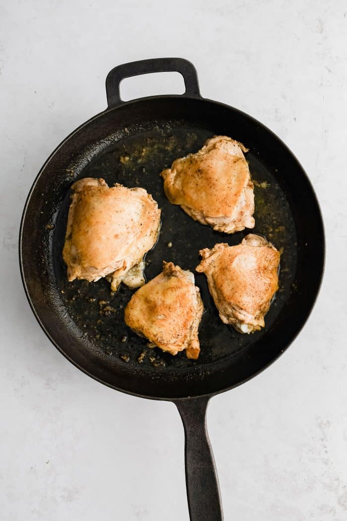 Four cooking bone-in skin-on marinated chicken thighs with seared golden skin in a large cast-iron skillet.