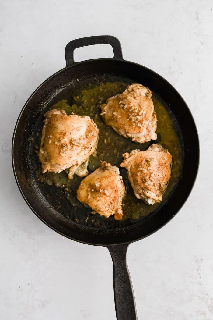 The remaining Greek lemon marinade added to a large cast iron skillet with four cooking bone-in skin-on marinated chicken thighs with seared golden skin.