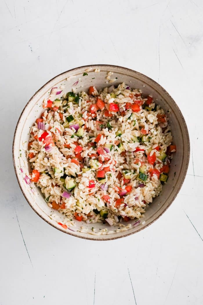 Large mixing bowl filled with cooked orzo, crumbled feta, chopped cucumber, tomatoes, red bell pepper, and red onions all tossed in a light lemon and parsley dressing.