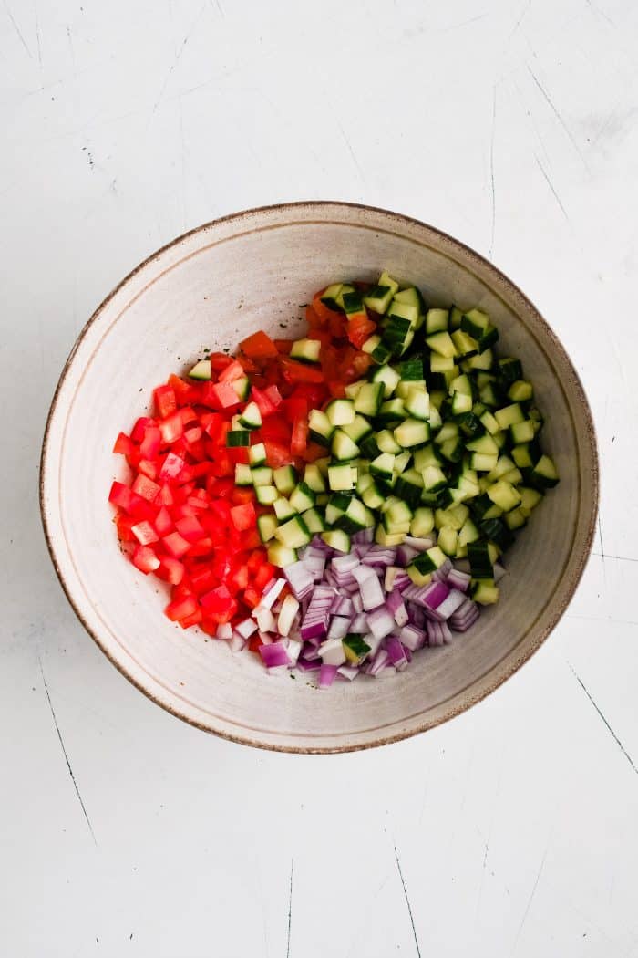 Large mixing bowl filled with diced cucumbers, red bell peppers, tomatoes, and red onion.