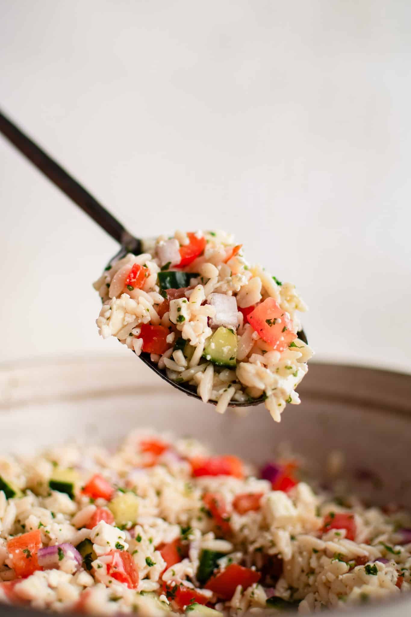 Large serving spoon filled with Mediterranean orzo salad hovering over a large bowl filled with the salad.